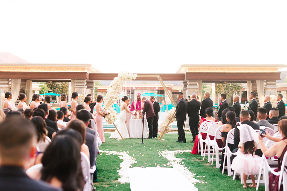  bride in a lace formfitting gown and a high neckline and the groom in a burgundy tuxedo with a black bow tie, and the bridesmaids in a rose blush gowns and the groomsmen in a black tuxedo with a black bow tie at the ceremony