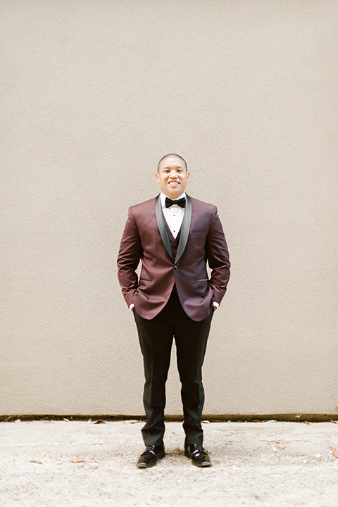  the groom in a burgundy tuxedo with a black bow tie smiling 