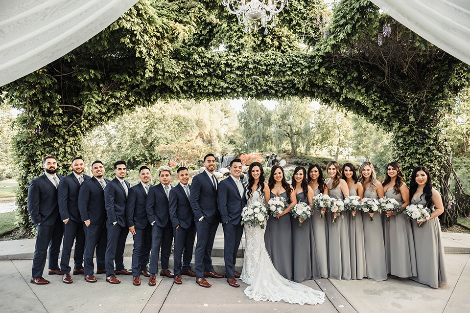  bride in a white formfitting gown and the groom in a navy suit with brown shoes and white long tie, the bridesmaids in light dusty sage gowns and groomsmen in navy blue suits and blue ties 