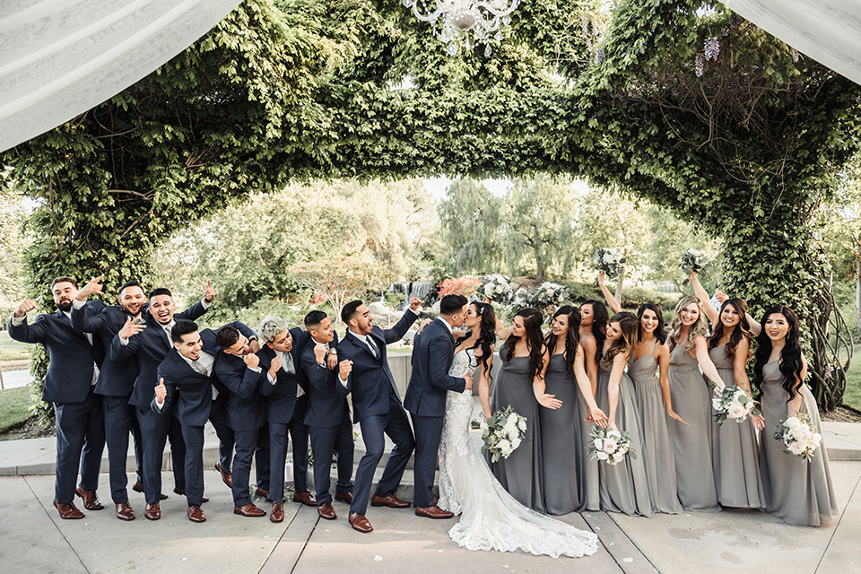  bride in a white formfitting gown and the groom in a navy suit with brown shoes and white long tie, the bridesmaids in light dusty sage gowns and groomsmen in navy blue suits and blue ties 