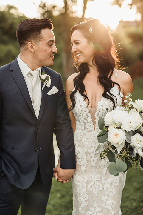  bride in a white lace gown with a crystal bodice and a deep v neckline and the groom in a navy notch lapel suit with a white long tie and brown shoes 
