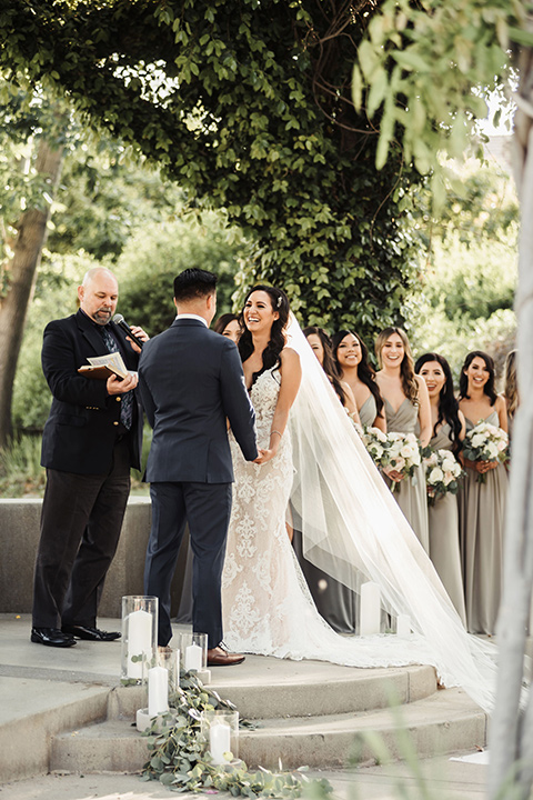 bride in a white lace gown with a crystal bodice and a deep v neckline and the groom in a navy notch lapel suit with a white long tie and brown shoes at the ceremony