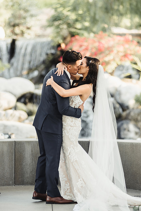  bride in a white lace gown with a crystal bodice and a deep v neckline and the groom in a navy notch lapel suit with a white long tie and brown shoes first kiss 