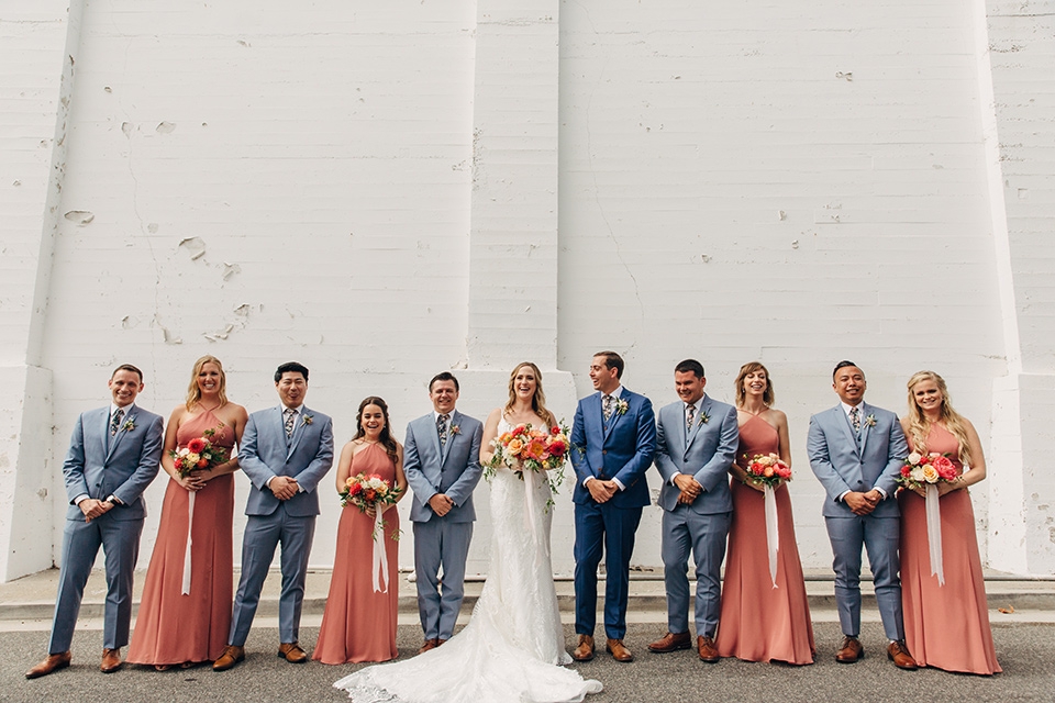  bride in a formfitting lace gown and the groom in a blue suit, the bridesmaids in desert sunset colored gowns, and the groomsmen in light blue suits