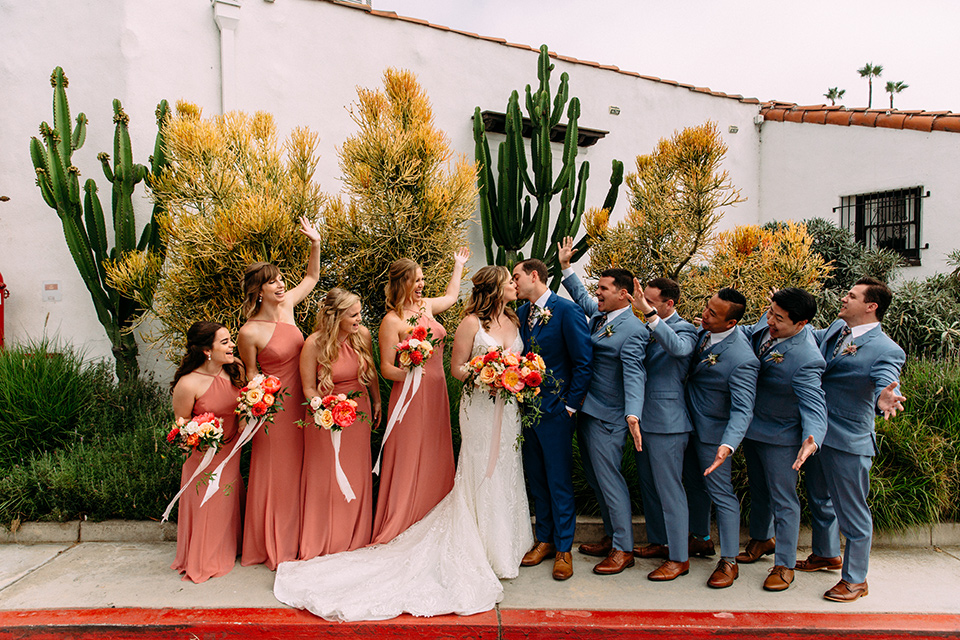  bride in a formfitting lace gown and the groom in a blue suit, the bridesmaids in desert sunset colored gowns, and the groomsmen in light blue suits