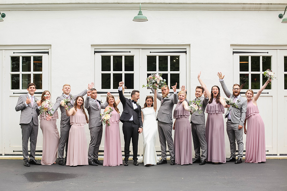  groom in a black tuxedo with a black bow tie and accessories bride in a white gown with a high neckline and veil, and the groomsmen in grey suits and bridesmaids in mauve and blush gowns