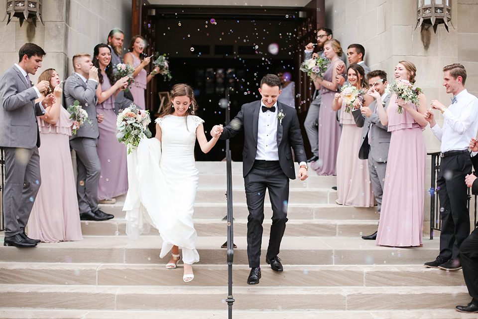  groom in a black tuxedo with a black bow tie and accessories bride in a white gown with a high neckline and veil, and the groomsmen in grey suits and bridesmaids in mauve and blush gowns