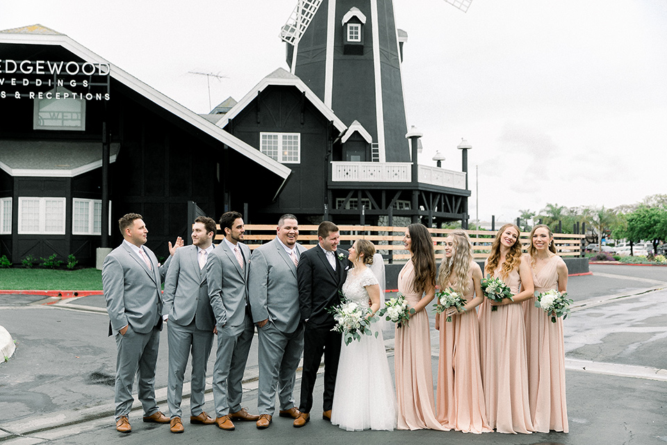  bride in a flowing gown with cap sleeves and the groom in an asphalt groom look, and the bridesmaids in peach gowns and the groomsmen in light grey suits
