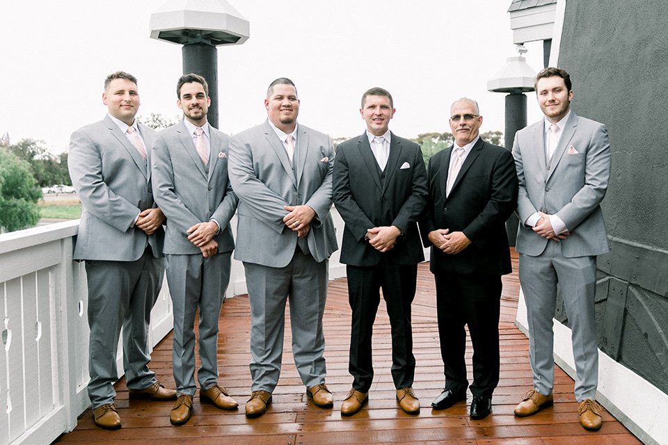  bride in a flowing gown with cap sleeves and the groom in an asphalt groom look, and the bridesmaids in peach gowns and the groomsmen in light grey suits