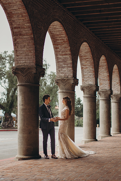  bride in a white lace gown with an illusion neckline and flowing train and the groom in a navy notch lapel suit with a deep teal long tie 