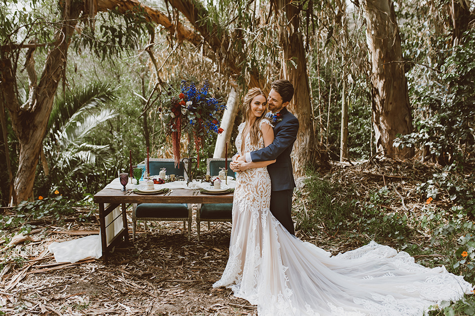  bride in a white lace gown with an illusion neckline and flowing train and the groom in a navy notch lapel suit with a deep teal long tie 