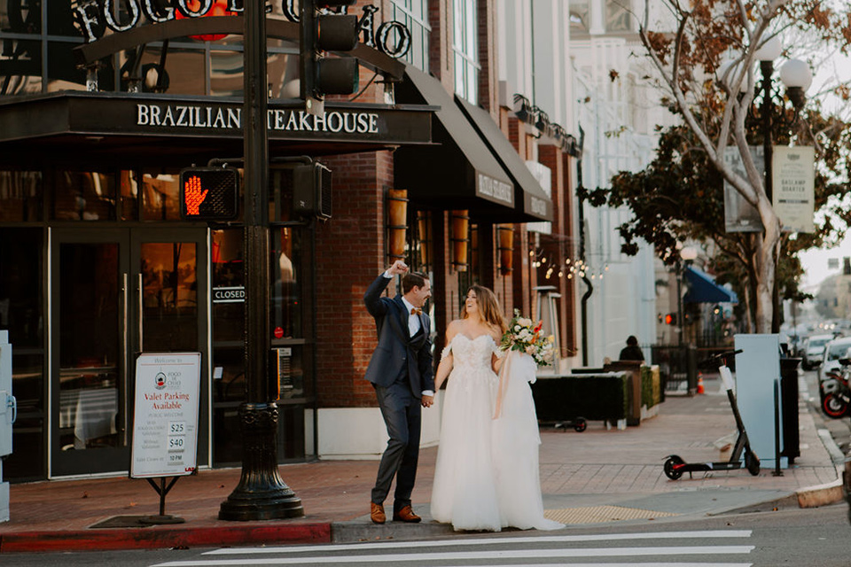  bride and groom crossing the street – bride in a full skirt gown with an off the shoulder detail and the groom in a dark blue suit with a bow tie