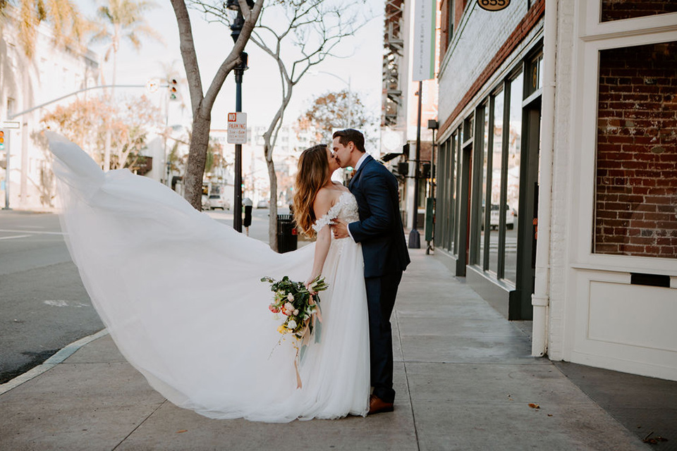  bride and groom crossing the street – bride in a full skirt gown with an off the shoulder detail and the groom in a dark blue suit with a bow tie