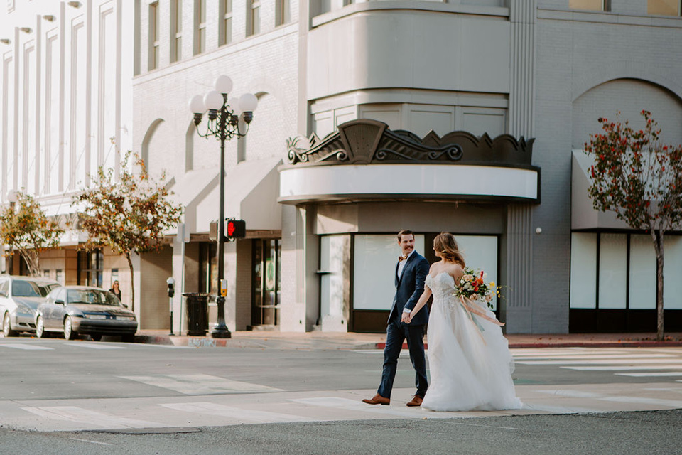  bride and groom crossing the street – bride in a full skirt gown with an off the shoulder detail and the groom in a dark blue suit with a bow tie