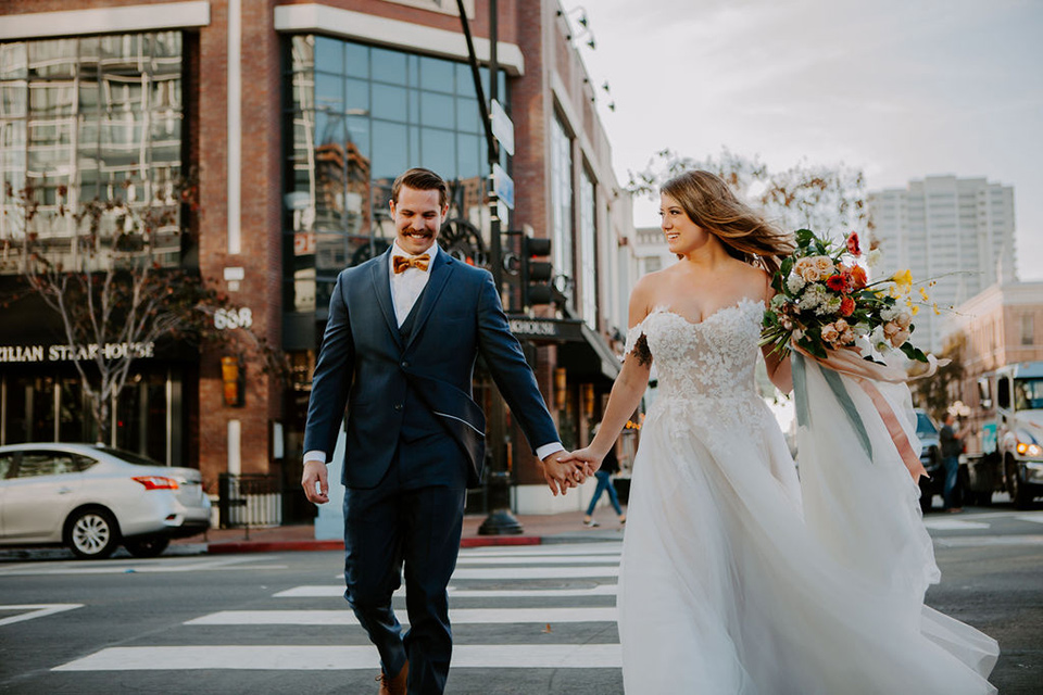  bride and groom crossing the street – bride in a full skirt gown with an off the shoulder detail and the groom in a dark blue suit with a bow tie