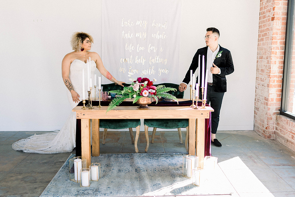  the bride is in a white formfitting lace gown with a sweetheart neckline and fun velvet shoes and the groom in a black velvet tuxedo with a cropped pant and bow tie at sweetheart table