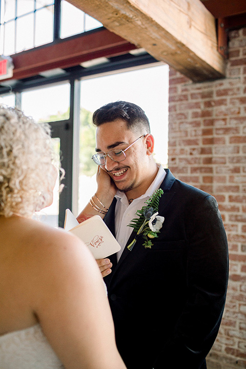  the bride is in a white formfitting lace gown with a sweetheart neckline and fun velvet shoes and the groom in a black velvet tuxedo with a cropped pant and bow tie together at the ceremony