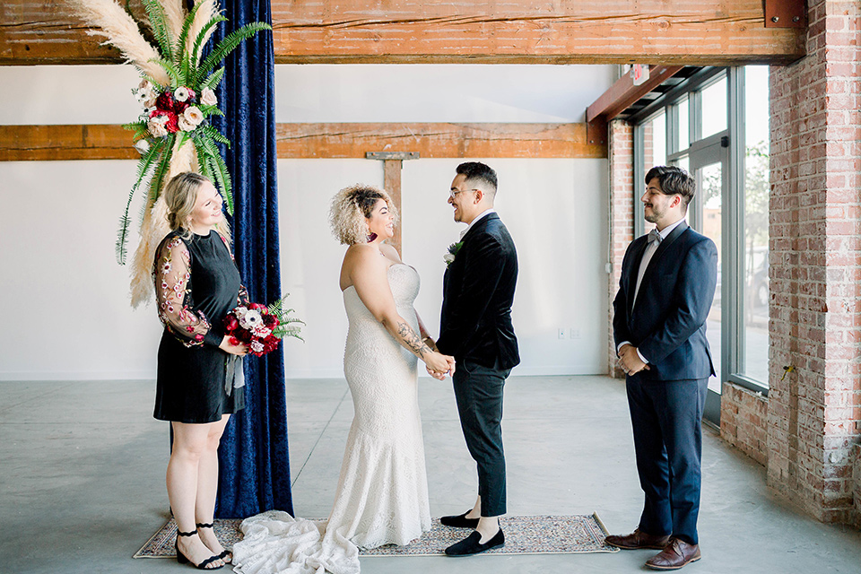  the bride is in a white formfitting lace gown with a sweetheart neckline and fun velvet shoes and the groom in a black velvet tuxedo with a cropped pant and bow tie at ceremony