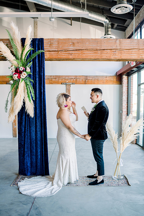  the bride is in a white formfitting lace gown with a sweetheart neckline and fun velvet shoes and the groom in a black velvet tuxedo with a cropped pant and bow tie together at the ceremony 