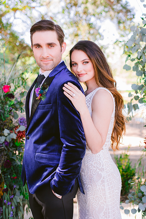  bride in a formfitting white gown with lace detailing and thin straps and the groom in a blue velvet tuxedo looking into the camera