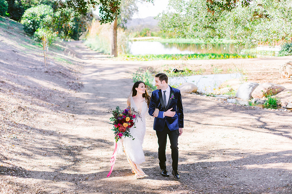 bride in a formfitting white gown with lace detailing and thin straps and the groom in a blue velvet tuxedo walking