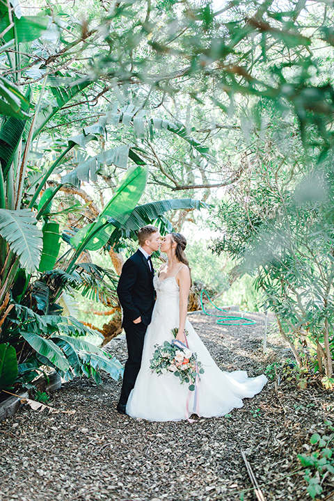  bride in a fit and flare gown with a strapless neckline and lace, the groom in a black tuxedo with a black bow tie standing under the trees