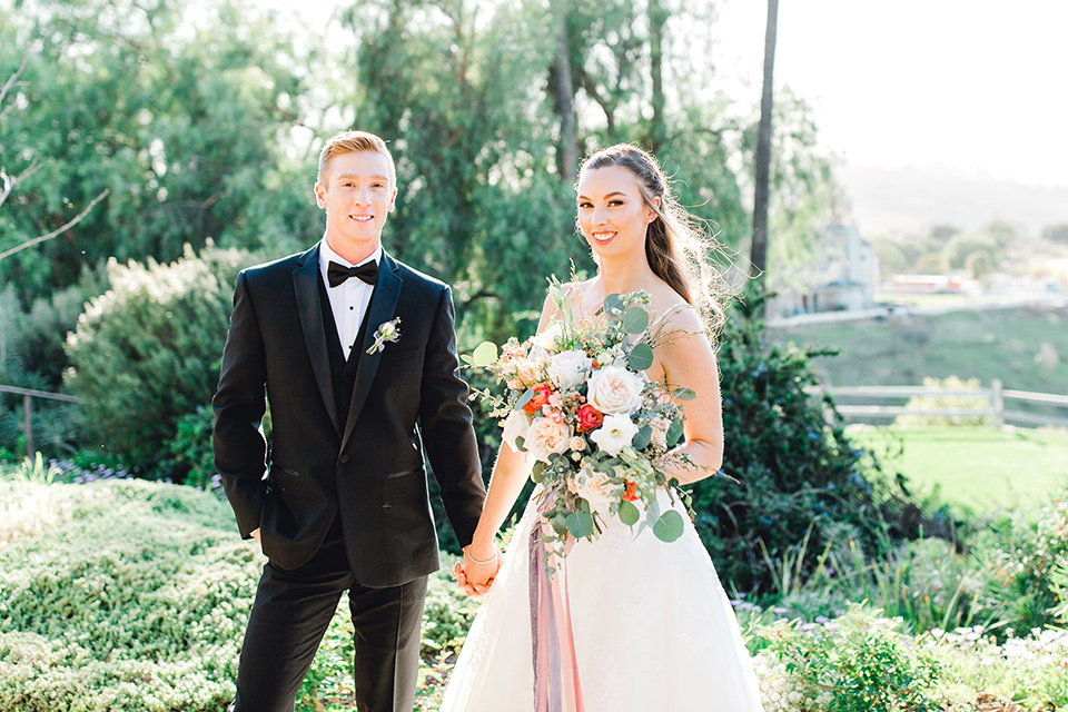  groom in a black tuxedo with a black bow tie and the bride in a white lace fit and flare gown looking at the camera