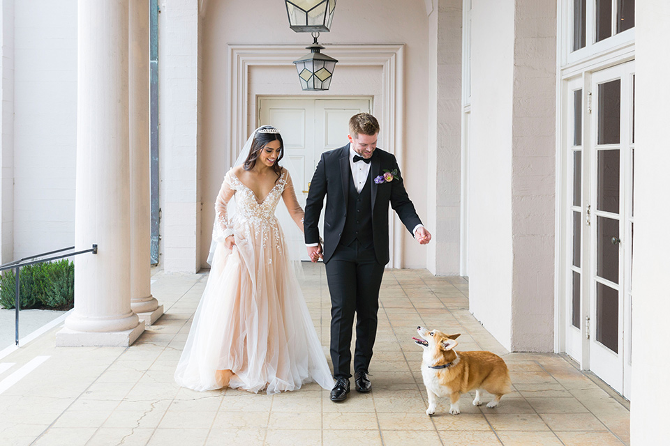  bride in a white lace ballgown with a sweetheart neckline and a regal looking crown and the groom in a black tuxedo with a black bow tie and pocket square walking their corgis