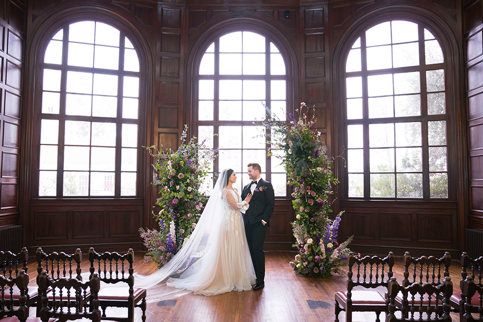  bride in a white lace ballgown with a sweetheart neckline and a regal looking crown and the groom in a black tuxedo with a black bow tie and pocket square at ceremony