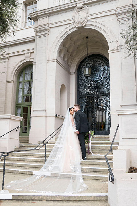  bride in a white lace ballgown with a sweetheart neckline and a regal looking crown and the groom in a black tuxedo with a black bow tie and pocket square on the steps 