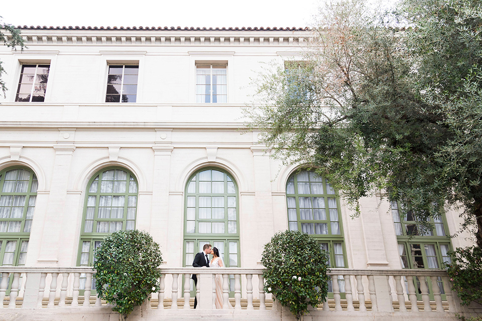  bride in a white lace ballgown with a sweetheart neckline and a regal looking crown and the groom in a black tuxedo with a black bow tie and pocket square in front of historic venue