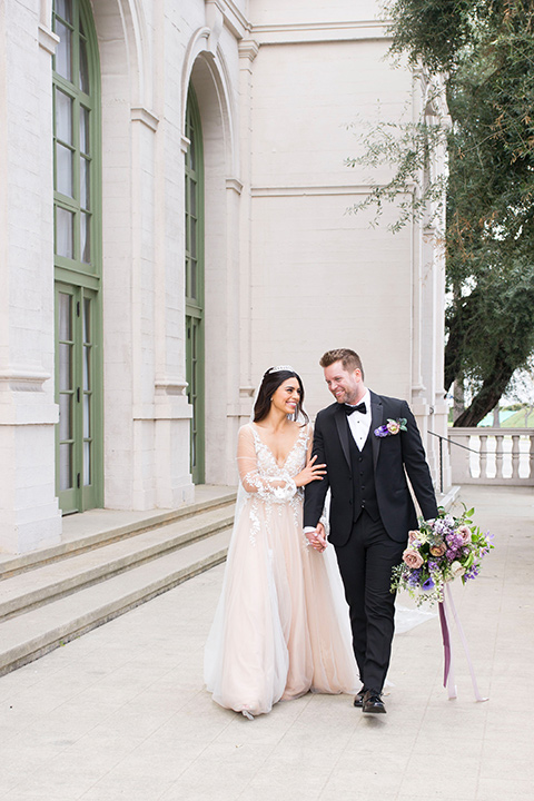  bride in a white lace ballgown with a sweetheart neckline and a regal looking crown and the groom in a black tuxedo with a black bow tie and pocket square on the steps 