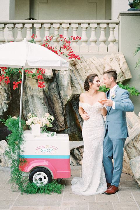  bride in a form fitting gown with an off the shoulder detail and the groom in a light blue suit next to bar cart