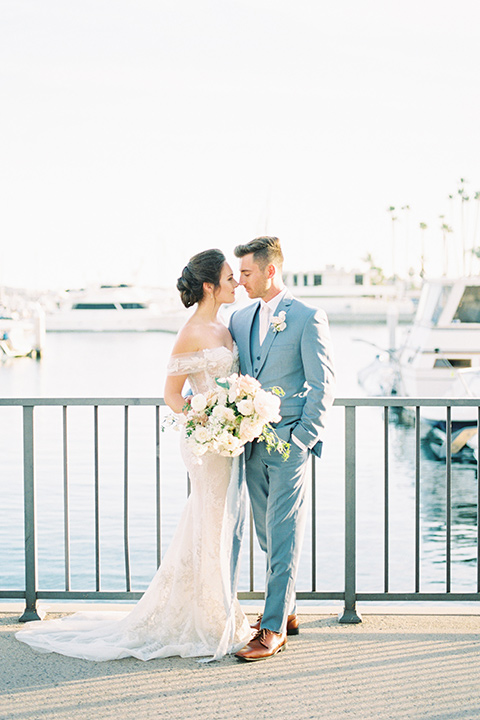  bride in a form fitting gown with an off the shoulder detail and the groom in a light blue suit by water 