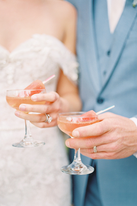  bride in a form fitting gown with an off the shoulder detail and the groom in a light blue suit next to bar cart close up on their drinks 