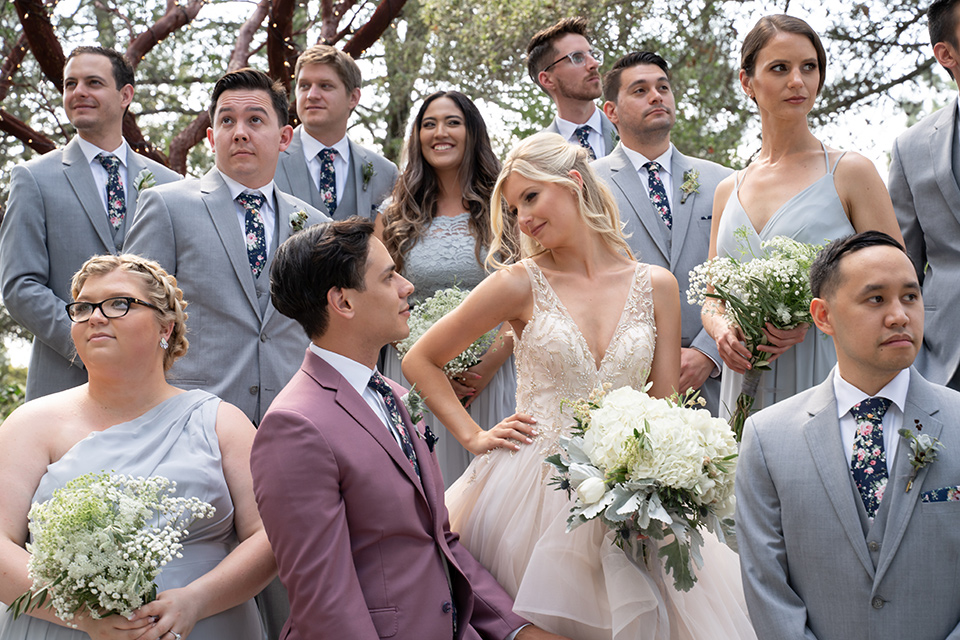  bride in a white ballgown with a deep-v neckline and crystal embellishments and the groom in a rose pink suit with a navy floral neck tie, and the bridesmaids in silver blue gowns and the groomsmen in heather grey suits with floral long ties 