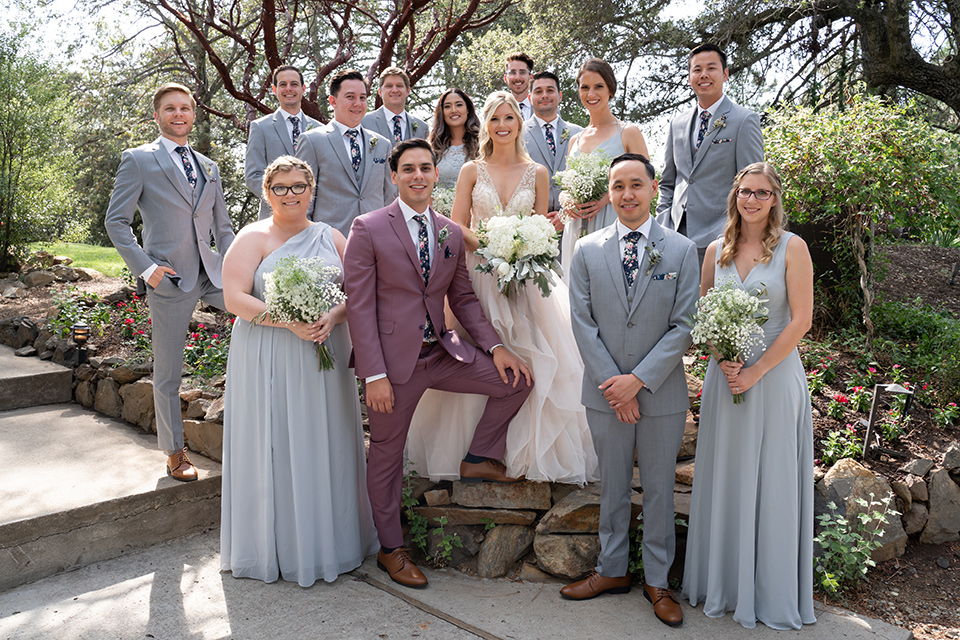  bride in a white ballgown with a deep-v neckline and crystal embellishments and the groom in a rose pink suit with a navy floral neck tie, and the bridesmaids in silver blue gowns and the groomsmen in heather grey suits with floral long ties 