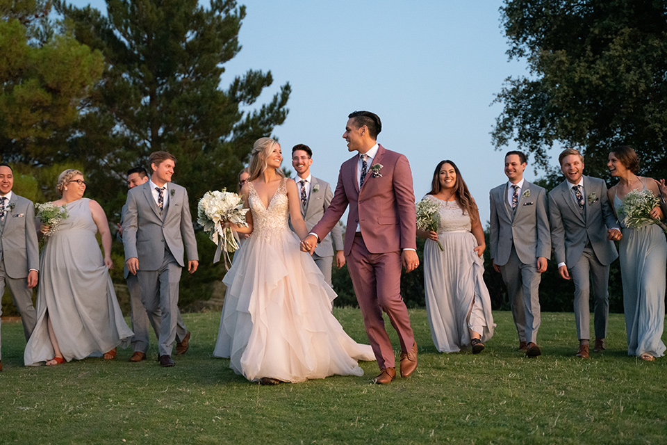  bride in a white ballgown with a deep-v neckline and crystal embellishments and the groom in a rose pink suit with a navy floral neck tie, and the bridesmaids in silver blue gowns and the groomsmen in heather grey suits with floral long ties 