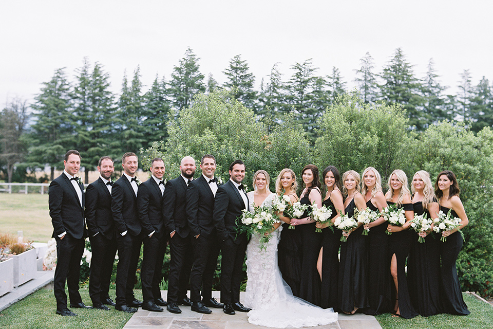  bride in a white gown with floral appliques and trumpet sleeves, the bridesmaids in black dresses in all different styles and the groom + groomsmen in black tuxedos and black bow ties