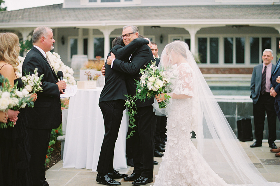  bride in a white gown with floral appliques and trumpet sleeves, the bridesmaids in black dresses in all different styles and the groom + groomsmen in black tuxedos and black bow ties