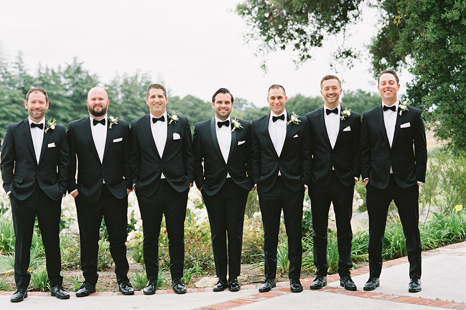  bride in a white gown with floral appliques and trumpet sleeves, the bridesmaids in black dresses in all different styles and the groom + groomsmen in black tuxedos and black bow ties
