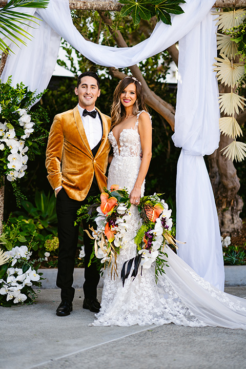 bride in a formfitting white lace gown with a sweetheart neckline and bold bouquet and the groom in a gold velvet tuxedo with black pants and black bow tie
