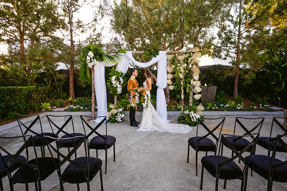  bride in a formfitting white lace gown with a sweetheart neckline and bold bouquet and the groom in a gold velvet tuxedo with black pants and black bow tie 