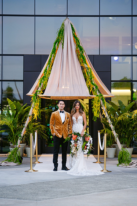  bride in a formfitting white lace gown with a sweetheart neckline and bold bouquet and the groom in a gold velvet tuxedo with black pants and black bow tie