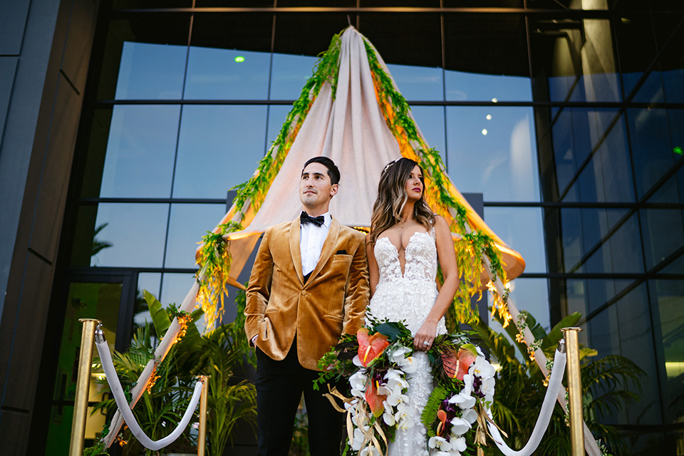  bride in a formfitting white lace gown with a sweetheart neckline and bold bouquet and the groom in a gold velvet tuxedo with black pants and black bow tie 