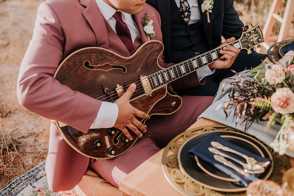  groom in a pink suit with a deep burgundy bow long tie and the other groom in a dark blue suit with a floral long tie at the table with a guitar