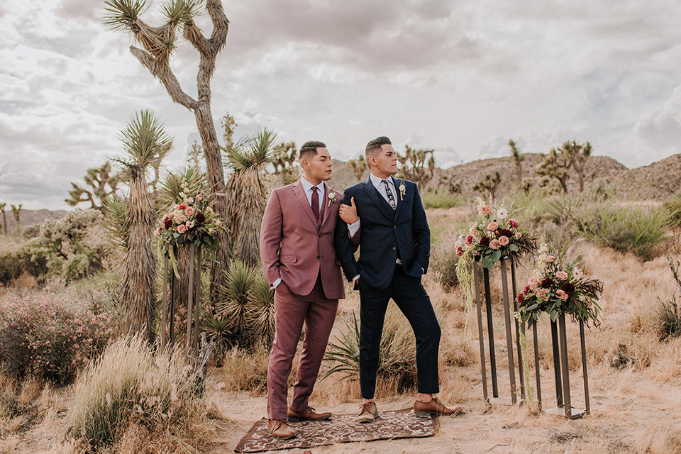  groom in a pink suit with a deep burgundy bow long tie and the other groom in a dark blue suit with a floral long tie