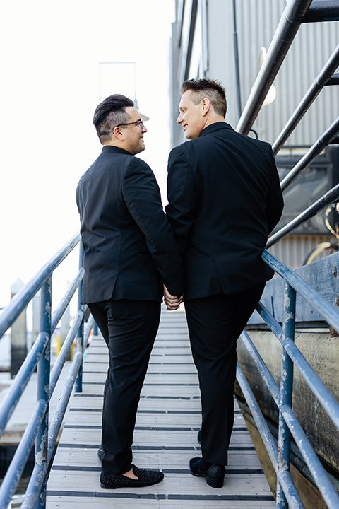  one groom in a black notch lapel tuxedo and black shirt, the other groom in a black shawl lapel tuxedo with a black shirt 