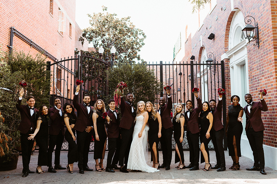 bride in a formfitting lace white gown with thin straps, and the groom in a burgundy tuxedo with a black bow tie.  The bridesmaids in black tea length gowns and the groomsmen in burgundy tuxedos