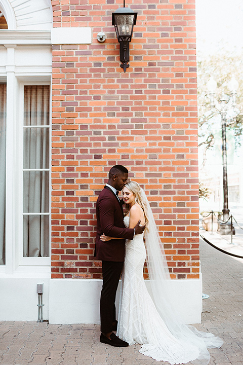 bride in a white formfitting gown with thin lace straps and cathedral veil and the groom in a burgundy tuxedo with a black bow tie